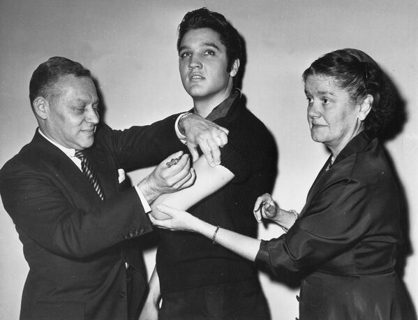 Elvis Presley recibe una vacuna contra la polio  en la ciudad de Nueva York, el 28 de octubre. Dr. Harold Fuerst de Nueva York se encarga de la inyección. La Dra. Leona Baumgartner, comisionada del departamento de salud de la Ciudad de Nueva York toma la mano del cantante. Presley es conocido por sus giros mientras presenta una canción. - Sputnik Mundo